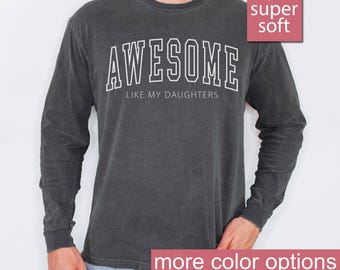 May include: A dark gray long-sleeve t-shirt with the text "AWESOME LIKE MY DAUGHTERS" printed in white. The shirt is made of a soft, comfortable fabric.