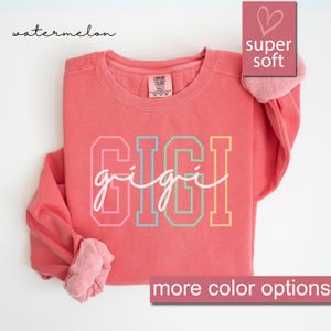 May include: A coral colored sweatshirt with the word "gigi" written in rainbow colors. The sweatshirt is made of a soft, comfortable fabric and has a crew neck.