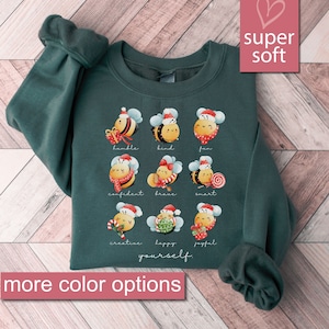 May include: A dark green sweatshirt featuring a Christmas bee design. The bees are wearing Santa hats and have different words written below them, including "humble", "kind", "fun", "confident", "brave", "smart", "creative", "happy", and "joyful". The text "yourself" is written below the bees. The sweatshirt is super soft.