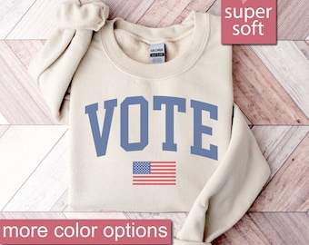 May include: A beige sweatshirt with the word "VOTE" in blue letters and an American flag underneath. The sweatshirt is on a wood background with the words "Sand", "super soft", and "more color options" in pink.