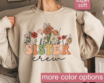 May include: Beige sweatshirt with a floral design and the words "SISTER crew" in colorful letters. The floral design includes orange, red, and yellow flowers. The text "super soft" and "more color options" are also visible.