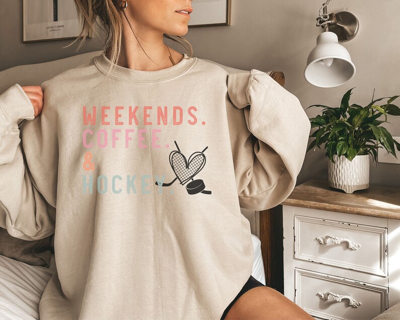 May include: A beige sweatshirt with the text "Weekends. Coffee & Hockey" in pink, blue, and black lettering. A heart with a hockey stick and puck is printed on the front.