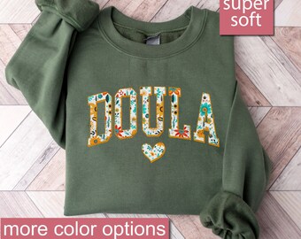 Może przedstawiać: Zielona bluza z nadrukiem słowa "DOULA" białymi literami z kwiatowym wzorem. Bluza jest super miękka i ma wzór serca pod słowem.