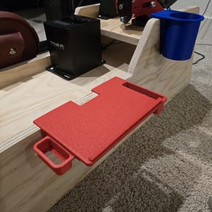 May include: A light-colored plywood racing simulator rig mount featuring a red 3D-printed rectangular tray with a small handle and a blue 3D-printed cup holder attached to the side. A black Moza Racing shifter is mounted on the wooden surface.
