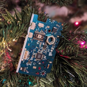 May include: A blue circuit board with a silver nut attached to it. The circuit board is hanging from a Christmas tree.