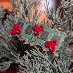 Op de afbeelding: Een unieke kerstversiering gemaakt van een groene printplaat, versierd met rode kralen. De versiering is gevormd als een hulstblad en rust op een bed van groene en zilveren kunstmatige dennentakken, met een dennenappel op de achtergrond.