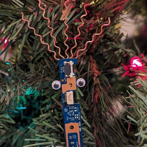 Puede incluir: Una placa de circuito azul con ojos saltones y pelo de alambre de cobre cuelga de un árbol de Navidad verde. La placa de circuito tiene el texto "W2-254 01121" impreso en ella.