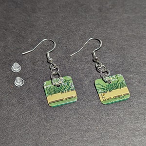 Puede incluir: Un par de pendientes de plata con dijes de estilo de placa de circuito verde. Los pendientes están sobre una superficie gris con dos respaldos de pendientes de plástico transparente.