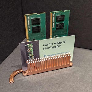 May include: A copper-colored metal business card holder with a decorative design. The holder is holding two green circuit boards and a white business card with the text "Cactus made of circuit parts? instagram.com/circuits.art".