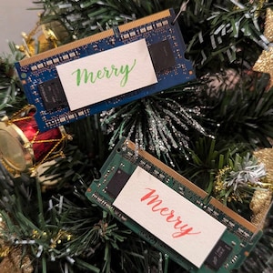 May include: Two computer memory sticks are decorated with white cards that say "Merry" in green and red calligraphy. The sticks are hanging on a green Christmas tree.