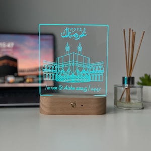 May include: A rectangular, illuminated LED night light with a wooden base. The light features a blue outline of a building and Arabic script. The text "Imran & Aisha 2025/1447" is also visible.