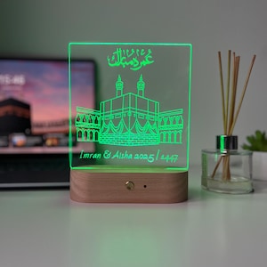 May include: A green-glowing acrylic LED lamp with a wooden base. The lamp features an etched illustration of a religious building and Arabic script. The text "Imran & Aisha 2025 / 1447" is also visible.