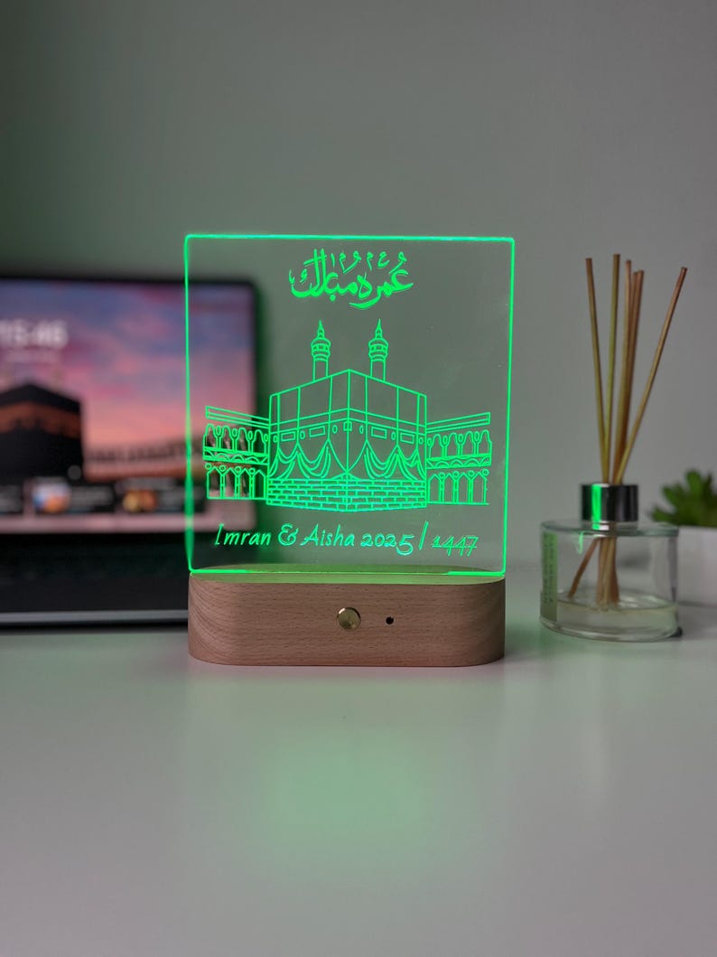 May include: A green-glowing acrylic LED lamp with a wooden base. The lamp features an etched illustration of a religious building and Arabic script. The text "Imran & Aisha 2025 / 1447" is also visible.