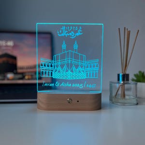 May include: A rectangular, illuminated LED night light with a wooden base. The light displays a detailed outline of a religious structure in blue, with Arabic script and the names "Imran & Aisha 2025 / 1447".