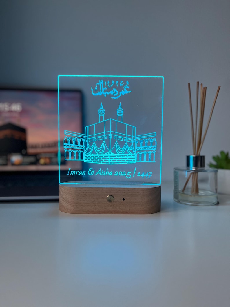 May include: A rectangular, illuminated LED night light with a wooden base. The light displays a detailed outline of a religious structure in blue, with Arabic script and the names "Imran & Aisha 2025 / 1447".