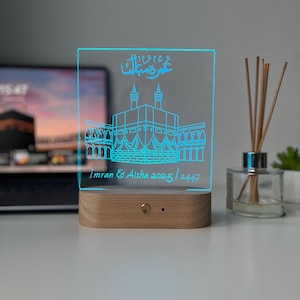 May include: A rectangular LED night light with a wooden base. The clear acrylic panel features a blue outline of a building with Arabic script above it. The names "Imran & Aisha" and the dates "2025 / 1447" are also displayed.
