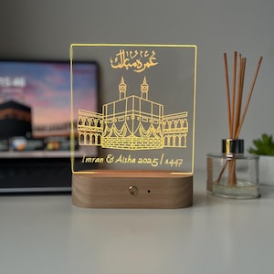 May include: A decorative LED night light featuring a detailed illustration of the Kaaba in Mecca, with Arabic calligraphy and the names "Imran & Aisha 2025/1447". The light has a wooden base and a clear acrylic panel.