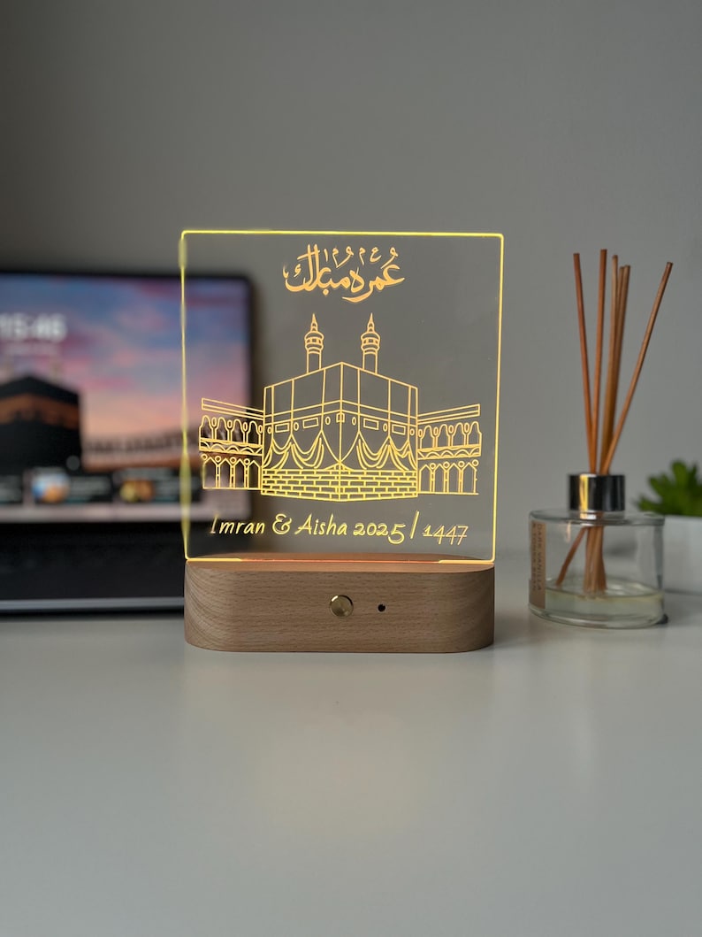 May include: A decorative LED night light featuring a detailed illustration of the Kaaba in Mecca, with Arabic calligraphy and the names "Imran & Aisha 2025/1447". The light has a wooden base and a clear acrylic panel.