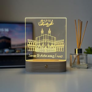 May include: A glowing LED night light with a rectangular acrylic panel featuring an etched illustration of a religious building. The base is made of wood. The text "Imran & Aisha 2025/1447" is visible.