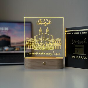 May include: A lighted acrylic plaque with a wooden base. The plaque features a gold-colored illustration of a religious structure and Arabic script. The names "Imran & Aisha" and the dates "2025/1447" are also inscribed. A book with the word "Umrah" is on the right.