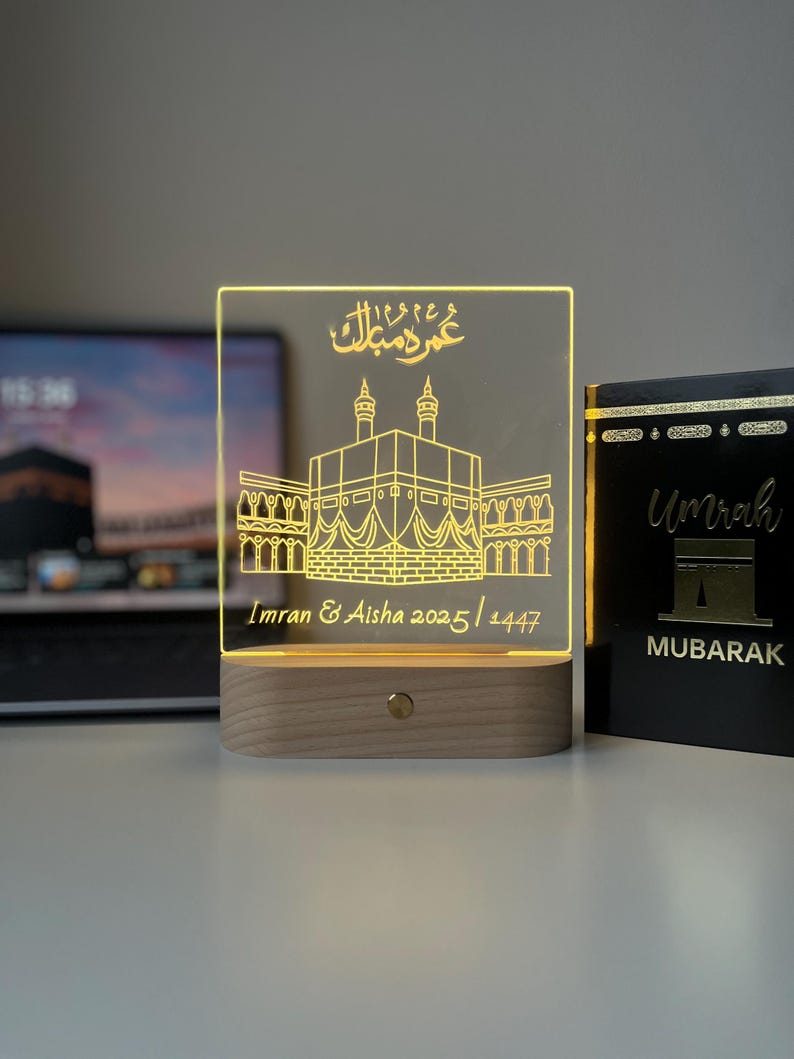 May include: A lighted acrylic plaque with a wooden base. The plaque features a gold-colored illustration of a religious structure and Arabic script. The names "Imran & Aisha" and the dates "2025/1447" are also inscribed. A book with the word "Umrah" is on the right.