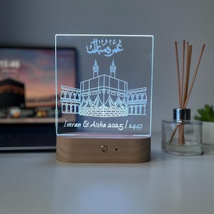 May include: A rectangular LED night light with a wooden base. The clear acrylic panel features an etched illustration of a religious building and Arabic script. The names "Imran & Aisha 2025 / 1447" are also etched on the panel.