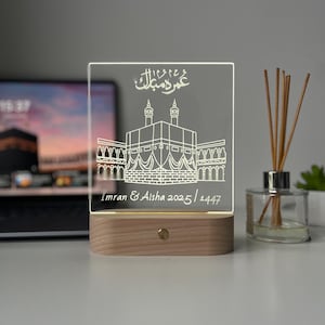 May include: A rectangular, illuminated acrylic plaque with a wooden base. The plaque features an etched illustration of a building with two minarets and Arabic script at the top. The names "Imran & Aisha 2025/1447" are inscribed below the building.