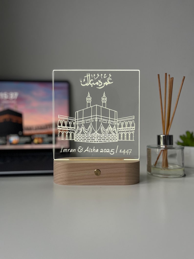May include: A rectangular, illuminated acrylic plaque with a wooden base. The plaque features an etched illustration of a building with two minarets and Arabic script at the top. The names "Imran & Aisha 2025/1447" are inscribed below the building.