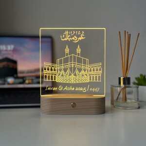 May include: A rectangular LED lamp with a wooden base. The clear acrylic panel features an etched illustration of a religious structure and Arabic script. The text "Imran & Aisha 2025/1447" is also etched on the panel.