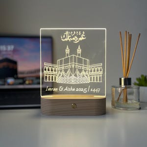 May include: A rectangular LED night light with a wooden base. The clear acrylic panel features an etched illustration of a religious structure, with Arabic script above and names and dates below. The light is on, illuminating the design.