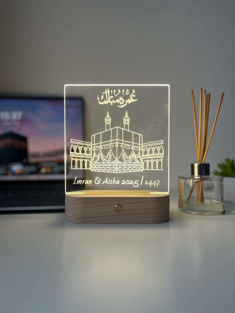 May include: A rectangular LED night light with a wooden base. The clear acrylic panel features an etched illustration of a religious structure, with Arabic script above and names and dates below. The light is on, illuminating the design.