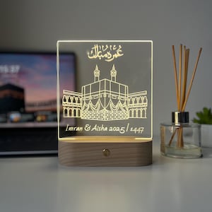 May include: A rectangular LED night light with a wooden base. The clear acrylic panel features an etched illustration of a religious building and Arabic text. The text "Imran & Aisha 2025/1447" is also etched on the panel.