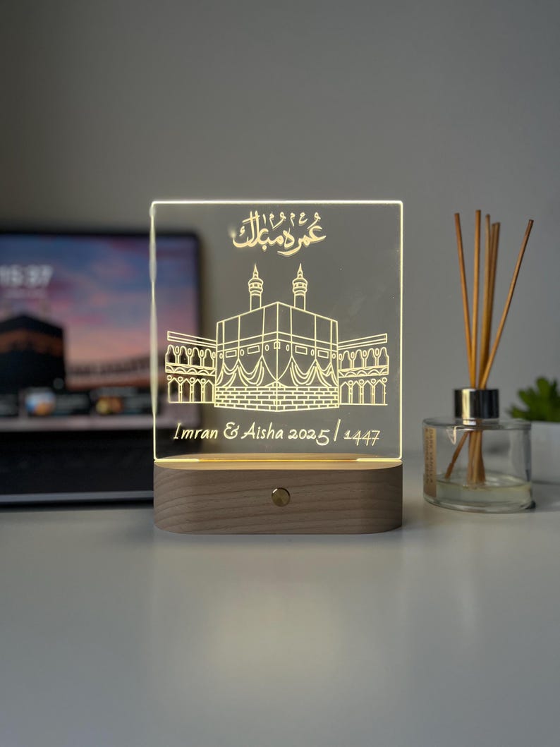 May include: A rectangular LED night light with a wooden base. The clear acrylic panel features an etched illustration of a religious building and Arabic text. The text "Imran & Aisha 2025/1447" is also etched on the panel.