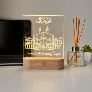 May include: A lighted acrylic night light with a wooden base. The design features an illustration of a religious building with Arabic script and the names "Imran & Aisha 2025 / 1447". The light is on, illuminating the design.