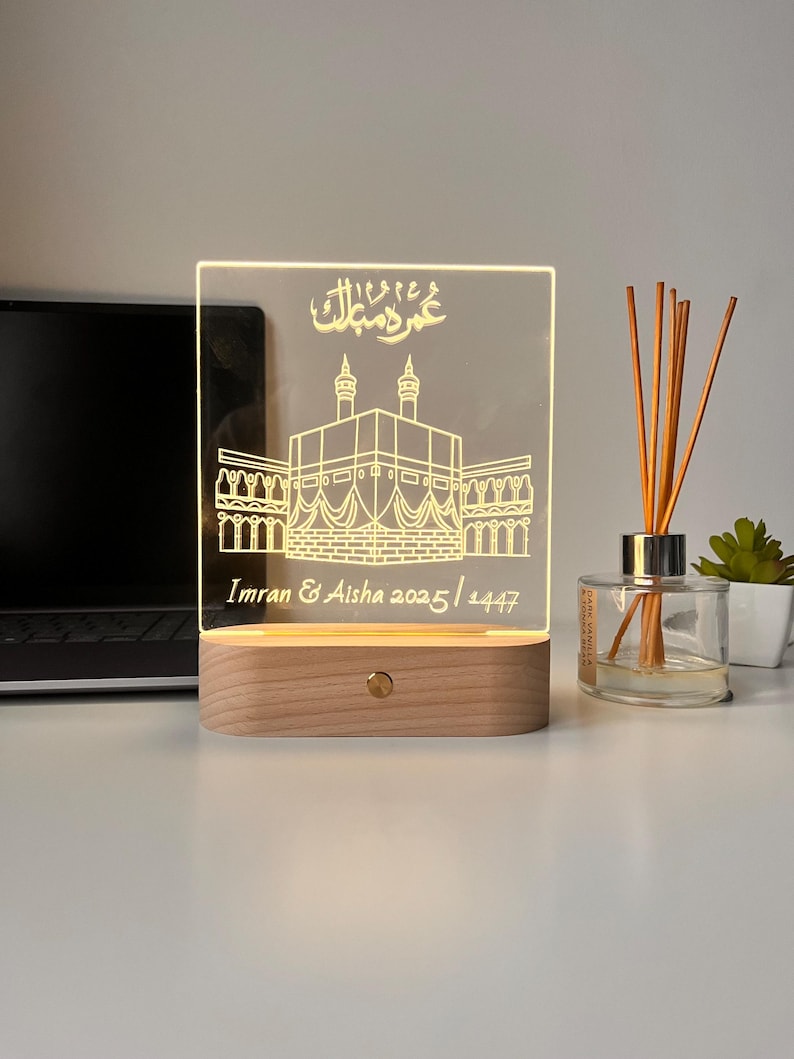 May include: A lighted acrylic night light with a wooden base. The design features an illustration of a religious building with Arabic script and the names "Imran & Aisha 2025 / 1447". The light is on, illuminating the design.