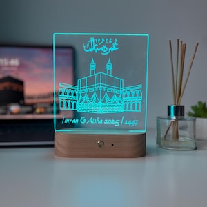 May include: A rectangular LED night light with a wooden base. The clear acrylic panel features a teal illustration of a building with Arabic script and the names "Imran & Aisha 2025 / 1447". The light is on, illuminating the design.