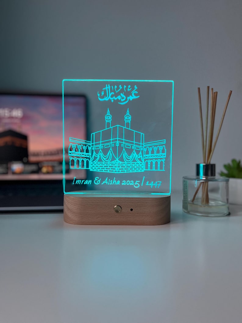 May include: A rectangular LED night light with a wooden base. The clear acrylic panel features a teal illustration of a building with Arabic script and the names "Imran & Aisha 2025 / 1447". The light is on, illuminating the design.