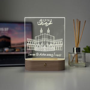 May include: A rectangular, illuminated acrylic plaque with a wooden base. The plaque features an etched illustration of a religious structure and Arabic script. The text "Imran & Aisha 2025/1447" is also etched on the plaque. The light is on.
