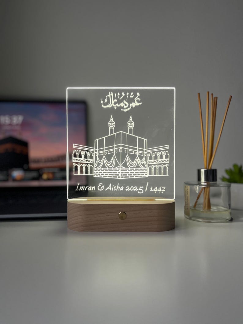May include: A rectangular, illuminated acrylic plaque with a wooden base. The plaque features an etched illustration of a religious structure and Arabic script. The text "Imran & Aisha 2025/1447" is also etched on the plaque. The light is on.