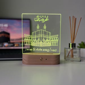May include: A personalized LED night light with a wooden base. The clear acrylic panel is etched with an illustration of a religious building and Arabic script. The names "Imran & Aisha" and the dates "2025/1447" are also etched.