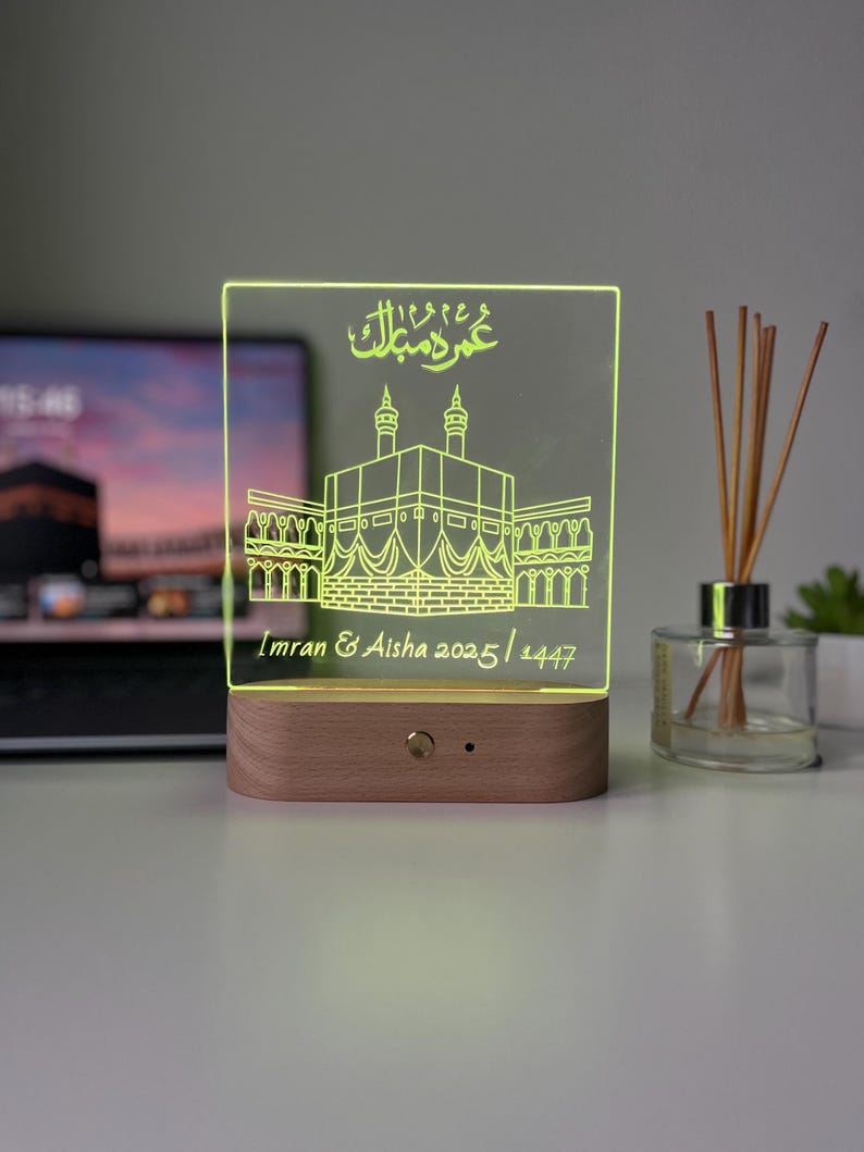 May include: A personalized LED night light with a wooden base. The clear acrylic panel is etched with an illustration of a religious building and Arabic script. The names "Imran & Aisha" and the dates "2025/1447" are also etched.