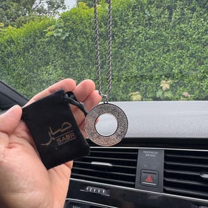 May include: A silver-toned circular pendant featuring intricate engraved Arabic calligraphy, suspended from a matching twisted rope chain. Beside the pendant is a small black velvet drawstring pouch with the text SABR COLLECTIONS.