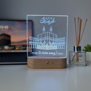 May include: A clear acrylic LED lamp with a wooden base. The lamp features an etched design of a building with Arabic script and the names "Imran & Aisha 2025/1447". The lamp is illuminated with a soft white light.