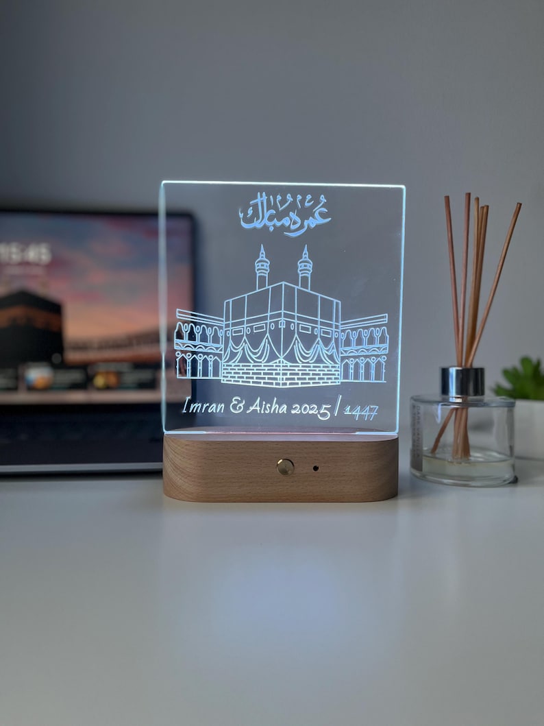 May include: A clear acrylic LED lamp with a wooden base. The lamp features an etched design of a building with Arabic script and the names "Imran & Aisha 2025/1447". The lamp is illuminated with a soft white light.