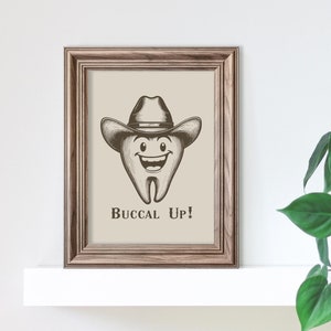 May include: A framed print with a cartoon tooth wearing a cowboy hat and the text "Buccal Up!" The print is on a white shelf with a green plant hanging over the edge.