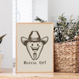 May include: A beige framed print with a cartoon tooth wearing a cowboy hat and the text "Buccal Up!" The tooth is smiling and has a happy expression.