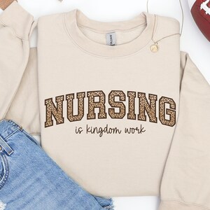 May include: A sand-colored sweatshirt with the word "NURSING" in a leopard print design, arched above the phrase "is kingdom work" in a simple font. The sweatshirt is folded with a pair of denim shorts and a gold necklace.
