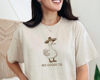 May include: A beige t-shirt with a brown illustration of a goose wearing a sombrero. The text "ME GOOSE-TA" is printed below the goose.