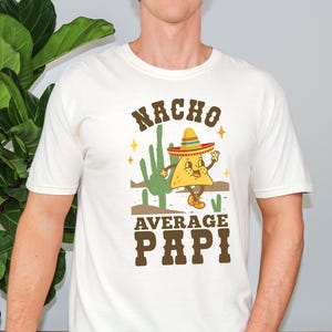 May include: Ivory-colored t-shirt with a graphic featuring a cartoon nacho wearing a sombrero, cactus, and the text "Nacho Average Papi". The design is in brown, yellow, and green.