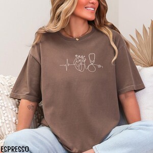 May include: A brown t-shirt with a white line drawing of a heart, heartbeat, and stethoscope. The design is centered on the front. The shirt has short sleeves and a relaxed fit. The word "ESPRESSO" is printed at the bottom of the image.